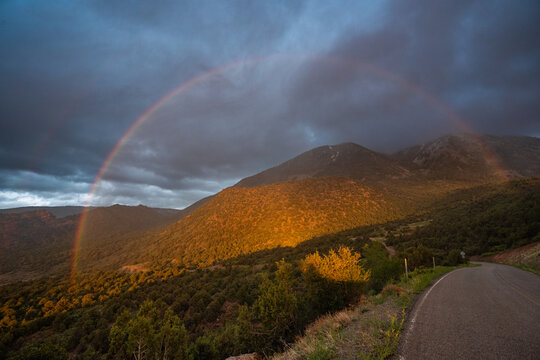 View of a bright double rainbow arching over the mountains with golden sunlight illuminating the slopes and a winding road in Moab, Utah, United States.
