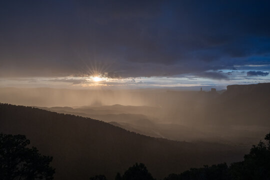 View of a sunburst breaking through dark storm clouds over a hazy desert valley with silhouetted hills in Moab, Utah, United States.