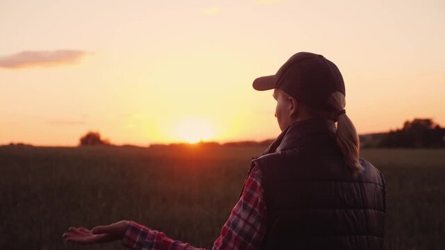 Female farmer wearing cap standing in wheat field at sunset and gesturing during phone conversation in countryside.