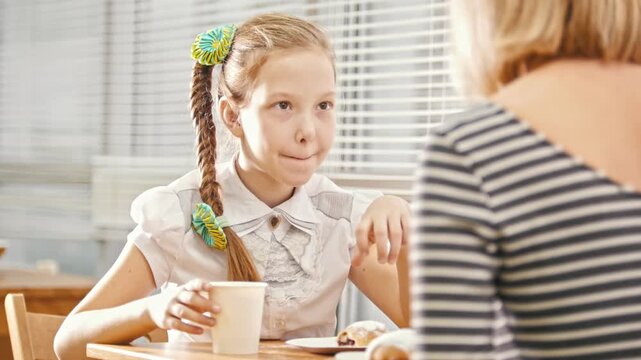 Girl with pigtail eats sweet cakes in the cafe, talks with her mum and licks her lips