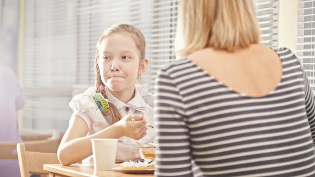 Girl with pigtail eats sweets in the cafe, talks with her mum and apes