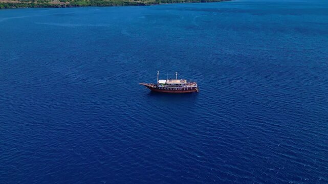 Traditional phinisi boat on open water with a person jumping off the bow into the sea. Magic aerial view descending panorama drone