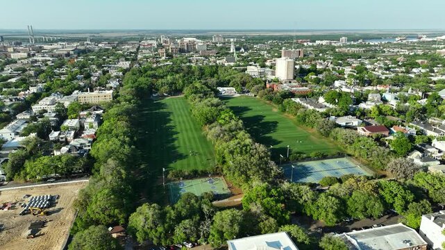Aerial drone view of Forsyth Park in Savannah, Georgia, showcasing lush green lawns, tree-lined paths, tennis courts, and the historic downtown skyline in the distance.