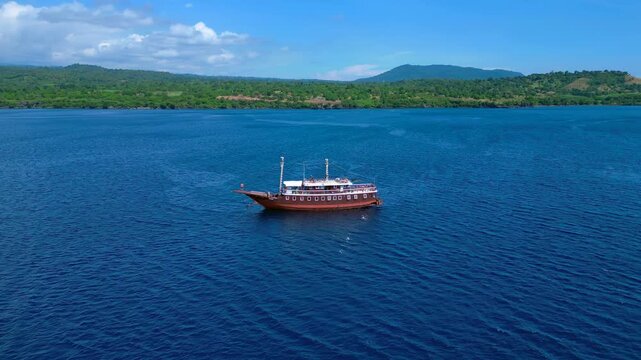 Traditional phinisi boat on open water with a person jumping off the bow into the sea. Great aerial view wide orbit panorama overview drone