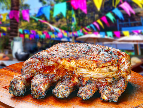 Roasted beef ribs on a wooden board at a Brazilian Festa Junina celebration, featuring colorful flags and festive outdoor background.