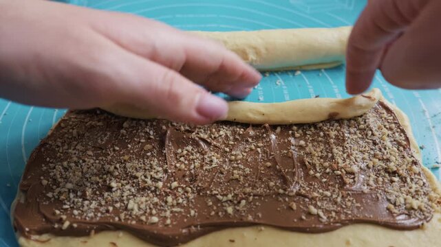 Female hands carefully roll sweet dough covered with thick chocolate spread and chopped nuts. Concept of domestic baking, homemade pastry preparation, and sweet artisan dessert making.