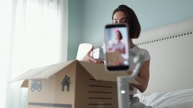 Young woman filming product unboxing with smartphone while opening cardboard package. Concept blogging, ecommerce and digital content.
