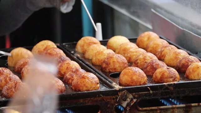Close-up of a street food vendor using a metal pick to flip golden-brown Takoyaki on a hot griddle.