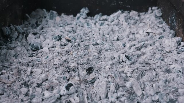 Burnt grey charcoal ashes and black coals rest inside a dark metal fire pit with crisp texture. Concept of outdoor cooking aftermath, fading heat, and authentic environmental destruction.