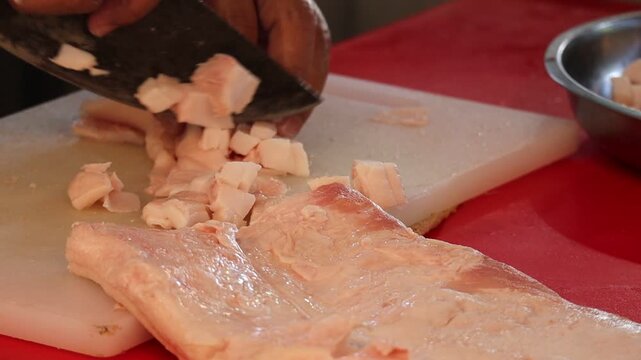 Close-up of a butcher or chef's hands using a large cleaver to dice raw pork fat on a white cutting board.