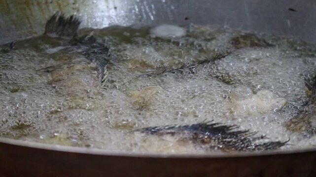 Close-up of whole snakeskin gourami fishes deep-frying in a large wok with bubbling hot oil.