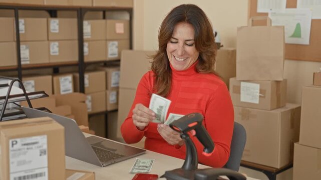 Woman counting cash at laptop while holding a barcode scanner in a packing building filled with parcels and stacked boxes; small business pride.