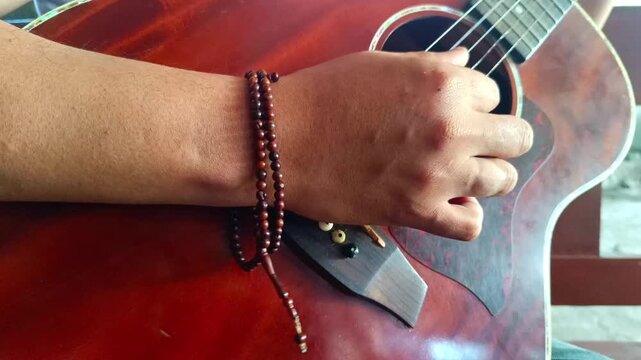 Footage of an Asian man's hand plucking the strings of an acoustic guitar while wearing a prayer bead bracelet accessory on his wrist.