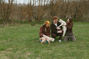 Naklejka premium Joyful family outdoors in a park shares smiles while playing with dogs and a young goat kid, capturing playful interaction, nature, and relaxed lifestyle. Natural light highlights warm mood, casual