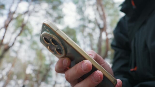 A person hand holds a modern green smartphone featuring a gold frame and large triple camera system against a blurred forest background. Concept of technology usage in outdoor environments.