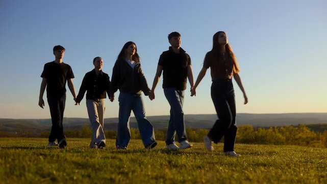 Five teenagers holding hands walking away on a grass hill at sunset