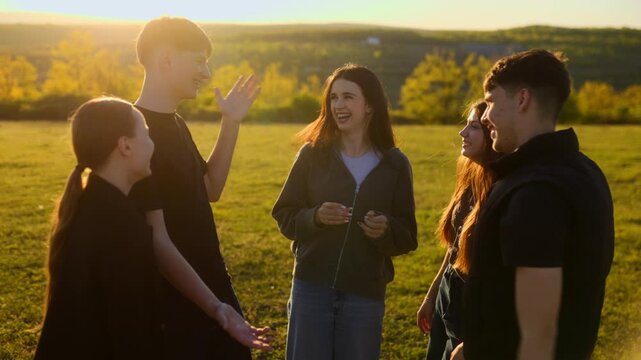 Five happy youth standing in a circle laughing on a field at sunset