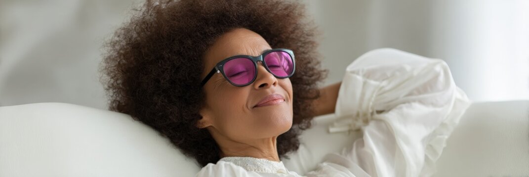 Relaxed female african adult reclining on a sofa wearing sunglasses smiling with eyes closed enjoying calm wellness and leisure at home in soft natural light