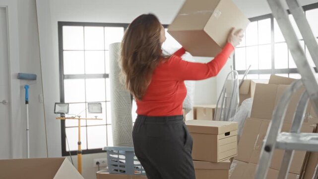 Woman holding cardboard box while lifting with both hands in building packed with moving supplies and ladder; happy moving transition.