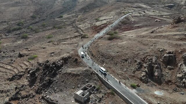 Aerial drone shot of white vans and people on the winding mountain road to Monte Verde, Sao Vicente. A high-angle view of the arid volcanic slopes and rural infrastructure in Cabo Verde
