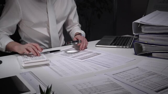 Person working late in an office, meticulously scrutinizing paperwork with a magnifying glass and pen, surrounded by a laptop, calculator, and binders filled with documents