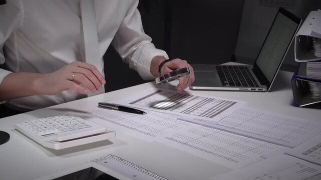 Person working late in an office, meticulously scrutinizing paperwork with a magnifying glass and pen, surrounded by a laptop, calculator, and binders filled with documents