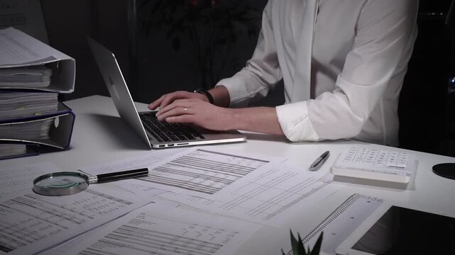 Dedicated finance professional at a cluttered desk, focused on analyzing documents and numbers with laptop, calculator and magnifying glass while managing company records and budgets