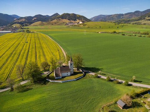 Aerial view of the Church of All Saints surrounded by vibrant yellow rapeseed and green fields with rolling mountains in the background Ludrova, Zilina Region, Slovakia.