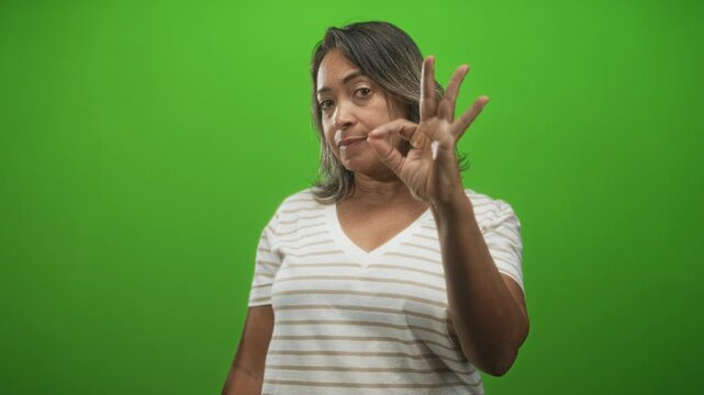 Hispanic woman wearing striped t shirt shows ok hand gesture with fingers near camera in green screen studio; confidence approval.