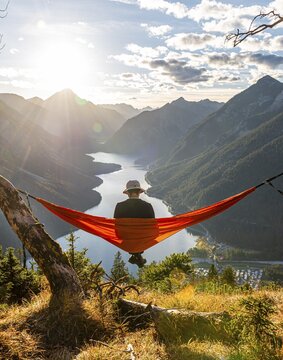 Man with a sun hat sitting in an orange hammock, panoramic view of mountains with lake, Plansee, Tyrol, Austria