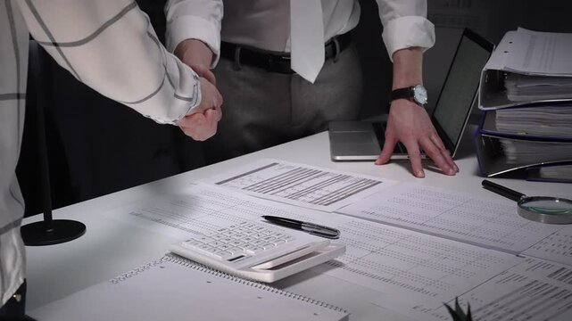 Business professionals shake hands over a cluttered desk of contracts, calculator and magnifying glass, sealing a trusted partnership after detailed financial review and late night work