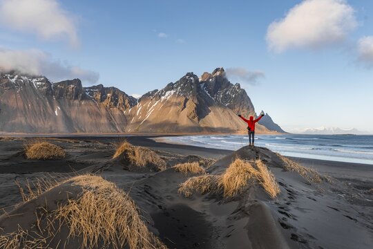 Man in red jacket stretches arms into the air, black lava beach, sandy beach, dunes with dry grass, mountains Klifatindur, Eystrahorn and Kambhorn, headland Stokksnes, mountain range Klifatindur, Austurland, East Iceland, Iceland