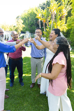 Diverse family and friends raising stemmed glasses, bride in white gown and veil standing on lawn