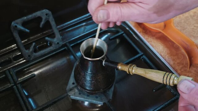 Hands stir ground coffee in a traditional copper cezve pot on a domestic gas stove burner. Concept of morning ritual, caffeine preparation, and artisanal brewing techniques.