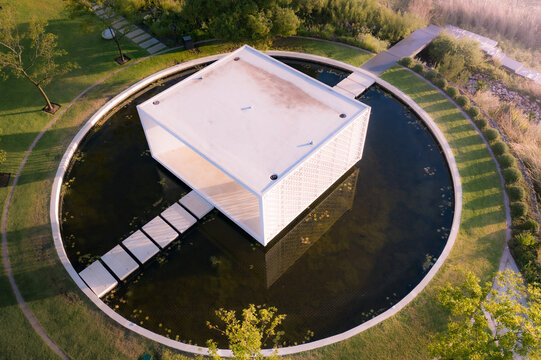 Aerial view of a modern white pavilion centered in a circular pond surrounded by lush green gardens and manicured lawns during golden hour in Cape Town, Western Cape, South Africa.