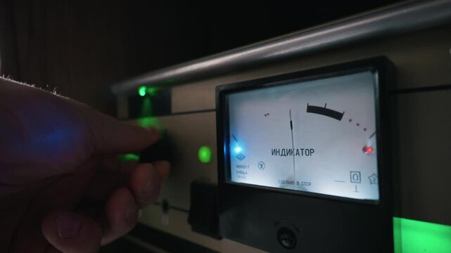 A person hand adjusts a control knob on a vintage tube audio amplifier device. The backlit analog panel measures sound decibel levels. The Russian word Indikator translates to Indicator.