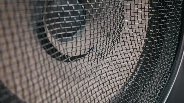 Large speaker cone moves rapidly back and forth behind a black metal wire mesh. Powerful vibrations demonstrate intense low frequency sound waves. Sharp focus highlights the rhythmic motion.