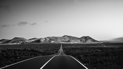 Black and white landscape photograph of an empty road leading through a dramatic volcanic landscape toward distant mountains. The image highlights strong leading lines, contrast, open space, and a sen © Erik