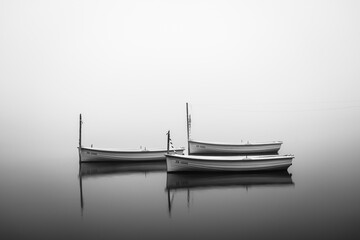 Black and white photo of small boats floating on a calm foggy lake with soft reflections and minimal atmosphere. © Erik