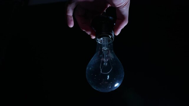 Male hands unscrew a brightly glowing incandescent light bulb from its socket. The warm tungsten light illuminates the fingers against a pitch-black background.