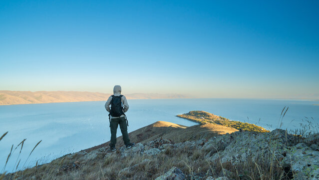 A hiker kneeling on a mountain slope above Lake Sevan, Armenia, admiring the calm blue water and distant hills under a clear morning sky.