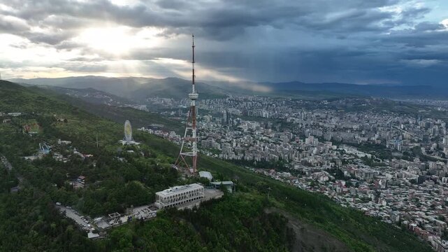 Aerial view of the Funicular Restaurant at Mtatsminda Park during sunset. Dynamic drone shot of a TV tower with Tbilisi city in the background.