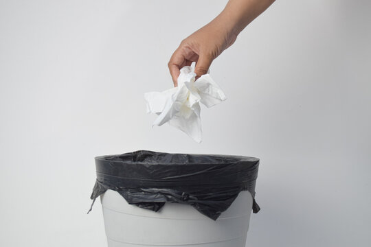Hand throwing crumpled white tissue paper into a trash bin with a black plastic liner against a white background.