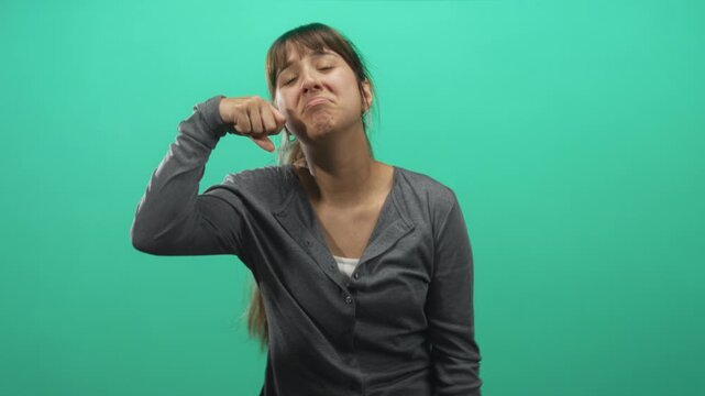 Woman in grey cardigan with long ponytail pinches her cheeks with fingers and makes a pouty smirk against a green screen in a studio; playful mischief.