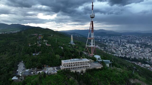 Aerial view of the Funicular Restaurant at Mtatsminda Park during sunset. Dynamic drone shot of a TV tower with Tbilisi city in the background.