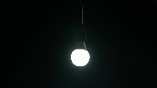 Hands carefully screw a glowing LED light bulb into a hanging electrical socket in total darkness. Concept of energy conservation, power outages, and bright new innovative ideas.
