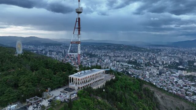 Aerial view of the Funicular Restaurant at Mtatsminda Park during sunset. Dynamic drone shot of a TV tower with Tbilisi city in the background.