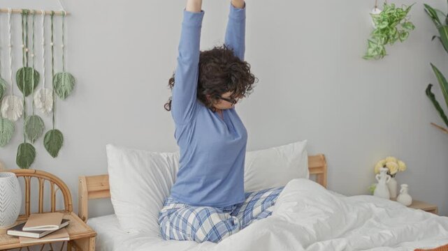 Woman stretching arms on white bed in a cozy building bedroom, wearing blue long sleeve pajamas and glasses while sitting on rumpled duvet; morning relaxation.