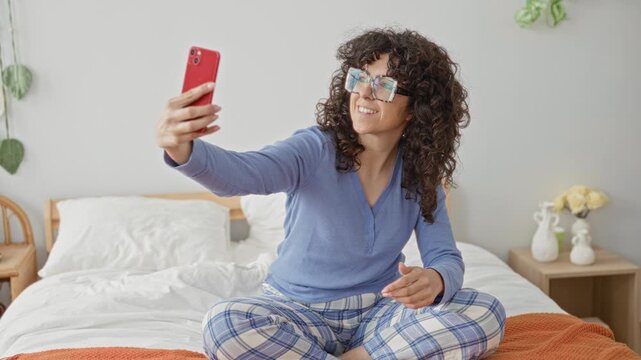Woman with curly hair and glasses holding smartphone and smiling while taking a selfie seated crosslegged on bed in building; serenity.