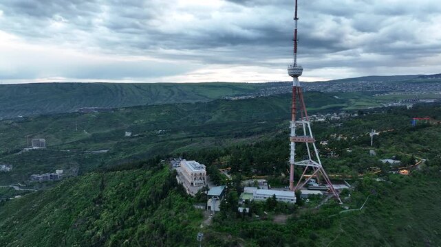 Aerial view of the Funicular Restaurant at Mtatsminda Park during sunset. Dynamic drone shot of a TV tower with Tbilisi city in the background.